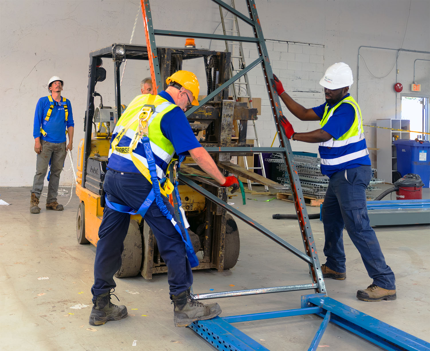 Two workers in safety gear maneuver a metal frame with a forklift nearby, while a third worker observes in a warehouse setting.